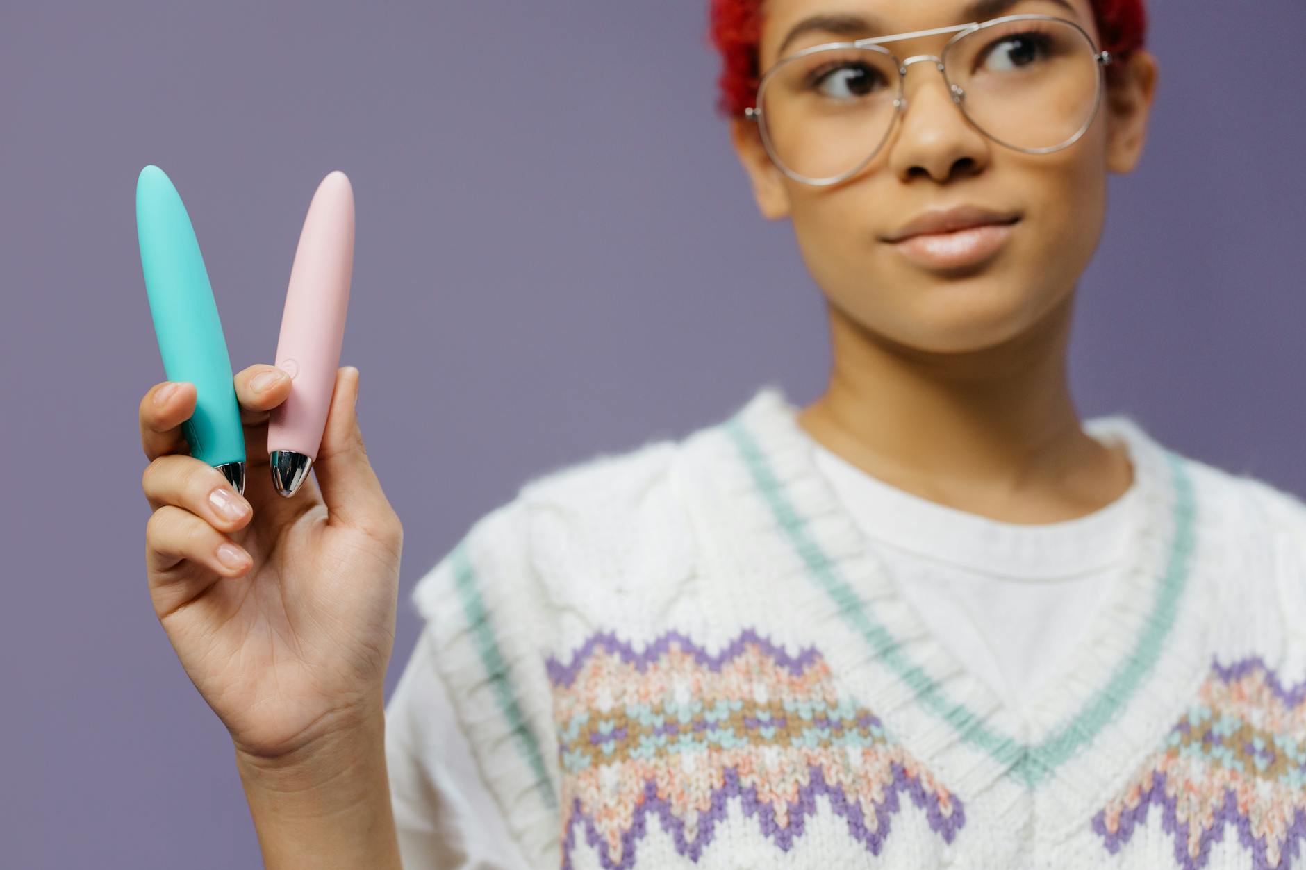 Woman with eyeglasses holding blue and pink silicone vibrators in a contemplative manner.