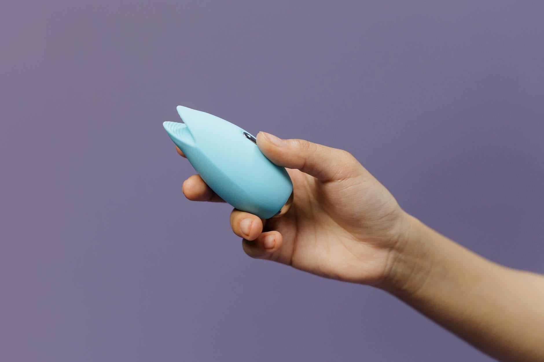 Detalhe íntimo de mãos segurando um vibrador azul cintilante contra um fundo roxo suave
